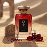 Bujairami Red Static Eau de Parfum 100ml bottle beside a carved stone in the sand with cherries and petals